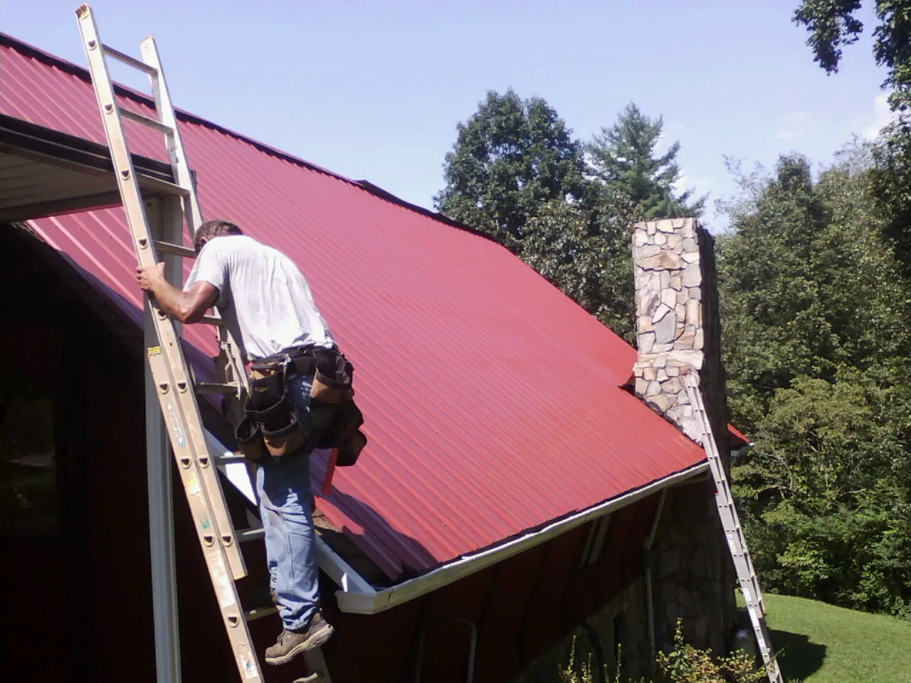 Roofer on a ladder beside a red metal roof and stone chimney — Soffit & Fascia Repair by Roofing Brothers in Reeseville